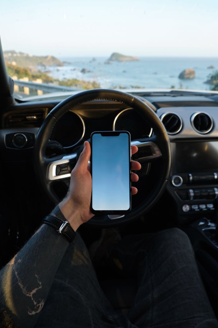 Services From above of crop unrecognizable tattooed male traveler using app on smartphone while sitting on driver seat in modern car against blurred seascape